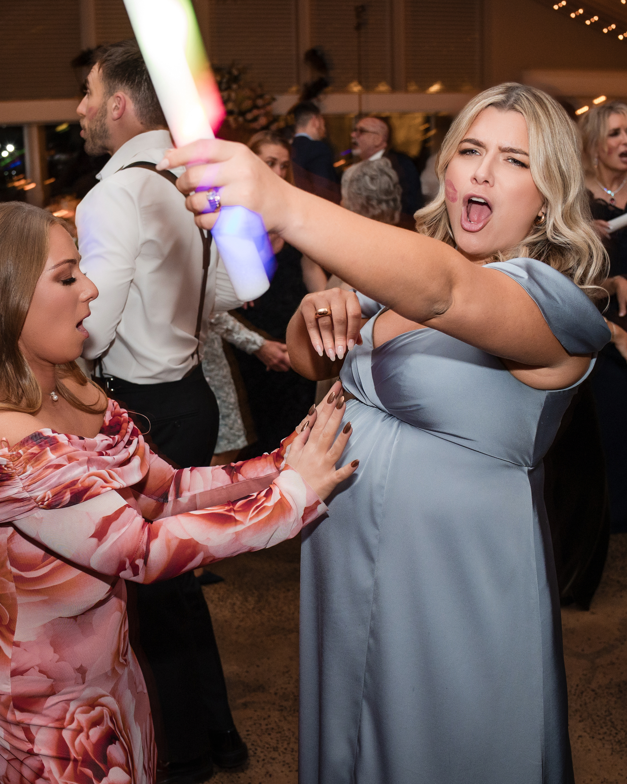 A woman in a blue dress holds a glowing stick and poses with an open mouth while another woman in a floral dress touches her baby bump at a lively Lake House Inn wedding.