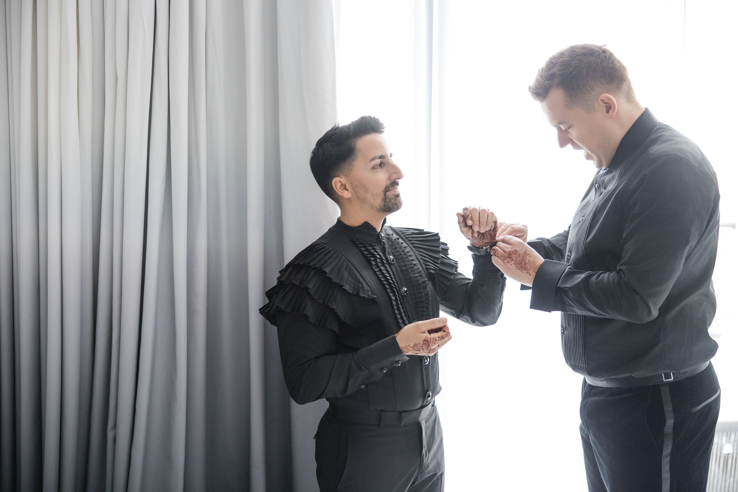 Two men dressed in formal black attire stand by a window at a House of Yes wedding, one helping the other adjust his cufflinks; both have intricate henna designs on their hands.