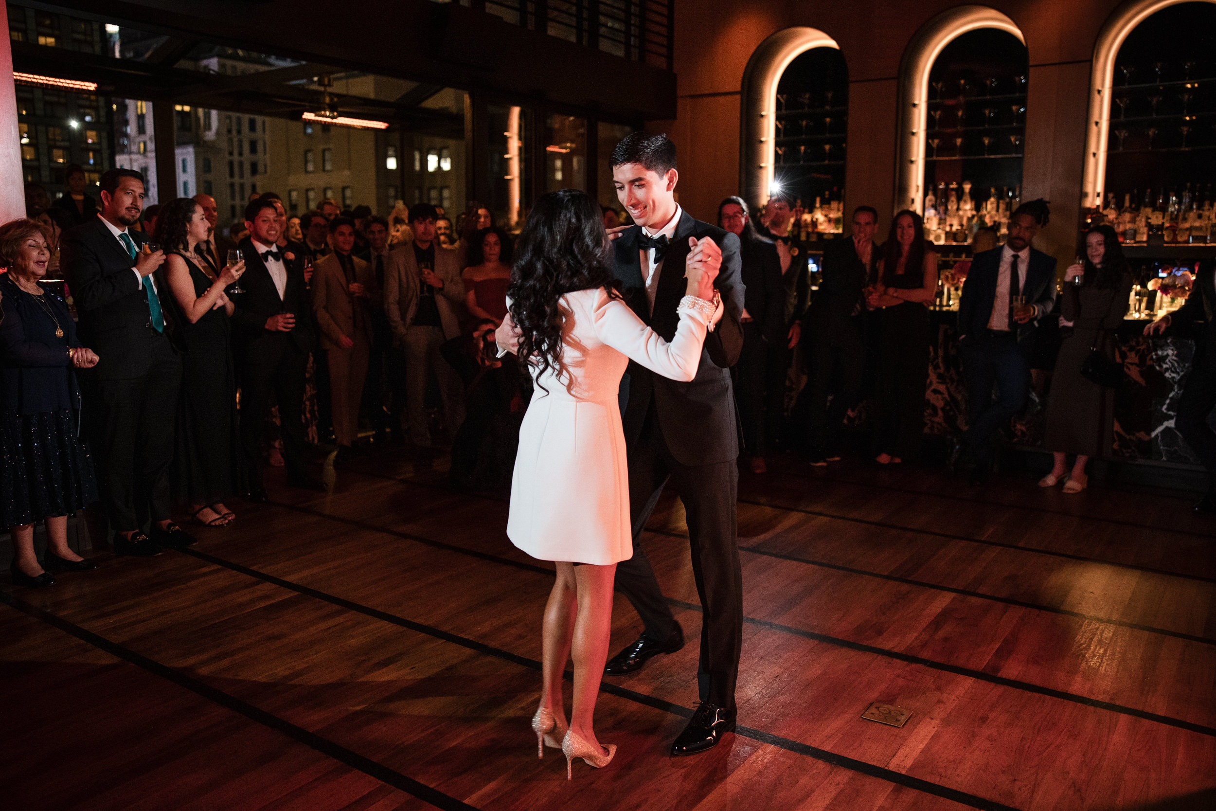A couple dances together in the center of a wooden floor at Castell Rooftop Lounge while a formally dressed crowd watches them in a dimly lit venue, celebrating their St. Patrick's Cathedral wedding.