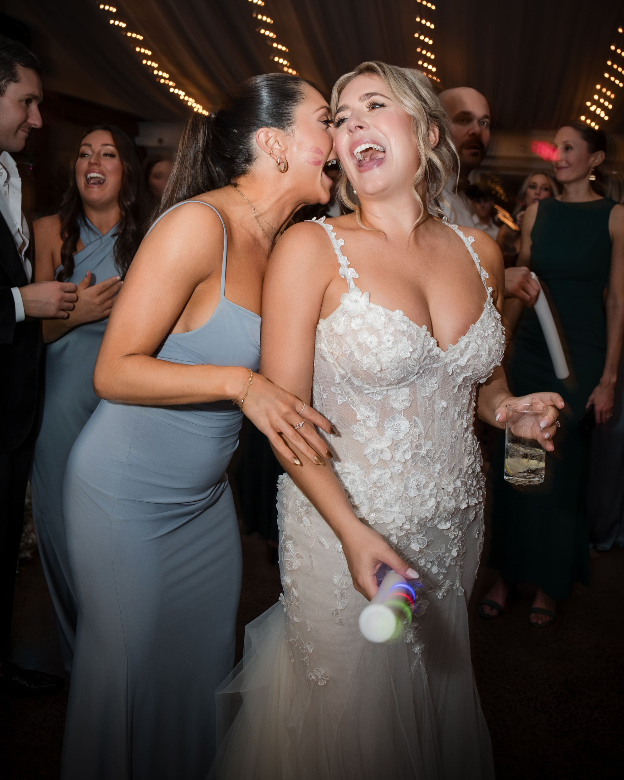 Two women in formal dresses laugh together at a Lake House Inn wedding reception; one holds a drink and a glow stick, while guests socialize in the background under string lights.