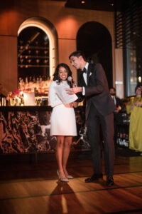 A woman in a white dress and a man in a black tuxedo stand together, smiling and holding a microphone on the wooden dance floor at Castell Rooftop Lounge, celebrating their stunning St. Patrick’s Cathedral wedding.
