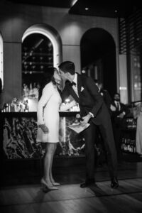 A woman in a white dress and a man in a suit kiss on the lips in the dimly lit Castell Rooftop Lounge, perhaps celebrating their St. Patrick’s Cathedral wedding, with people and a marble bar in the background.