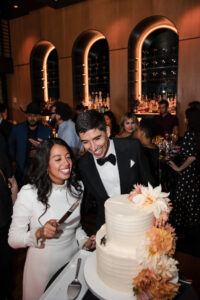 A couple dressed in formal attire smiles as they prepare to cut a white tiered wedding cake decorated with flowers at their Castell Rooftop Lounge reception, celebrating after a stunning St. Patrick’s Cathedral wedding surrounded by guests.