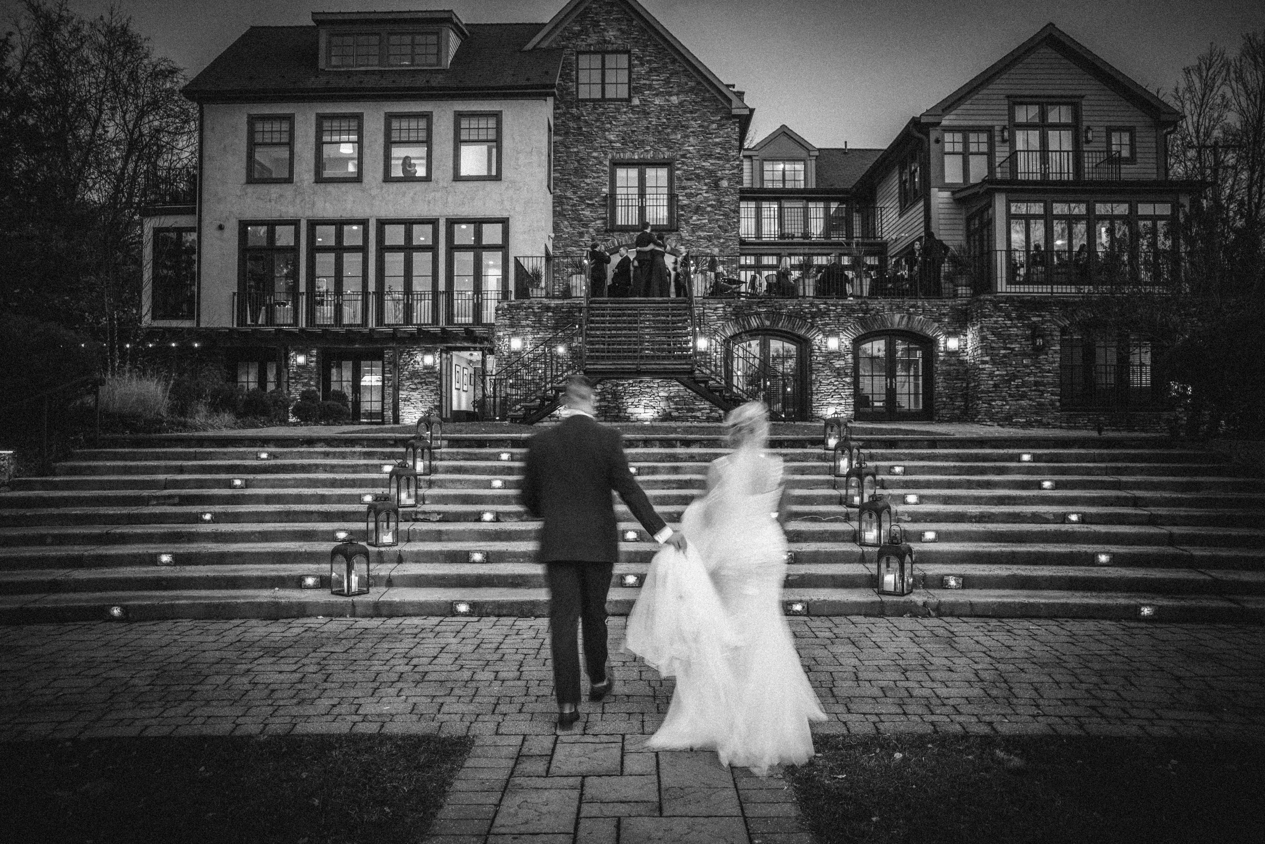A bride and groom walk up outdoor steps toward a large, stone-faced building at dusk during their Lake House Inn wedding, with guests standing on the balcony above.