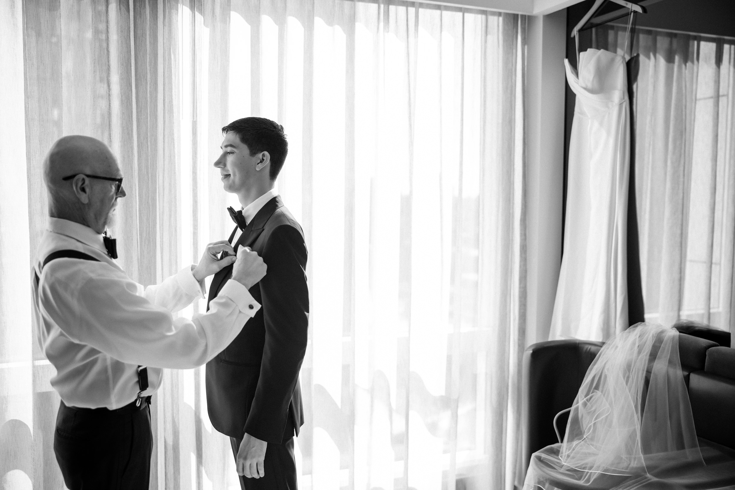A man helps another adjust his bowtie in a sunlit room with a wedding dress and veil hanging nearby, preparing for a St. Patrick's Cathedral wedding before celebrating at Castell Rooftop Lounge.