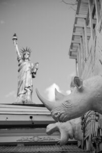 A black-and-white photo showing a statue of the Statue of Liberty in the background and a large rhinoceros head sculpture, reminiscent of a House of Yes wedding, mounted on a building in the foreground.