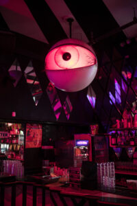 A bar interior with pink and purple lighting features a large, illuminated eyeball decoration hanging from the ceiling above the counter, evoking the vibrant atmosphere of a House of Yes wedding.