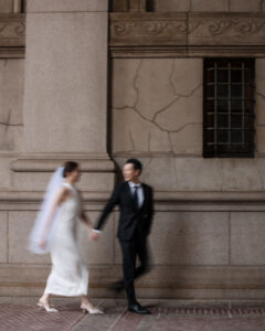 A bride in a white dress and veil holds hands with a groom in a black suit as they walk past a stone building, capturing timeless City Hall subway station wedding photos.