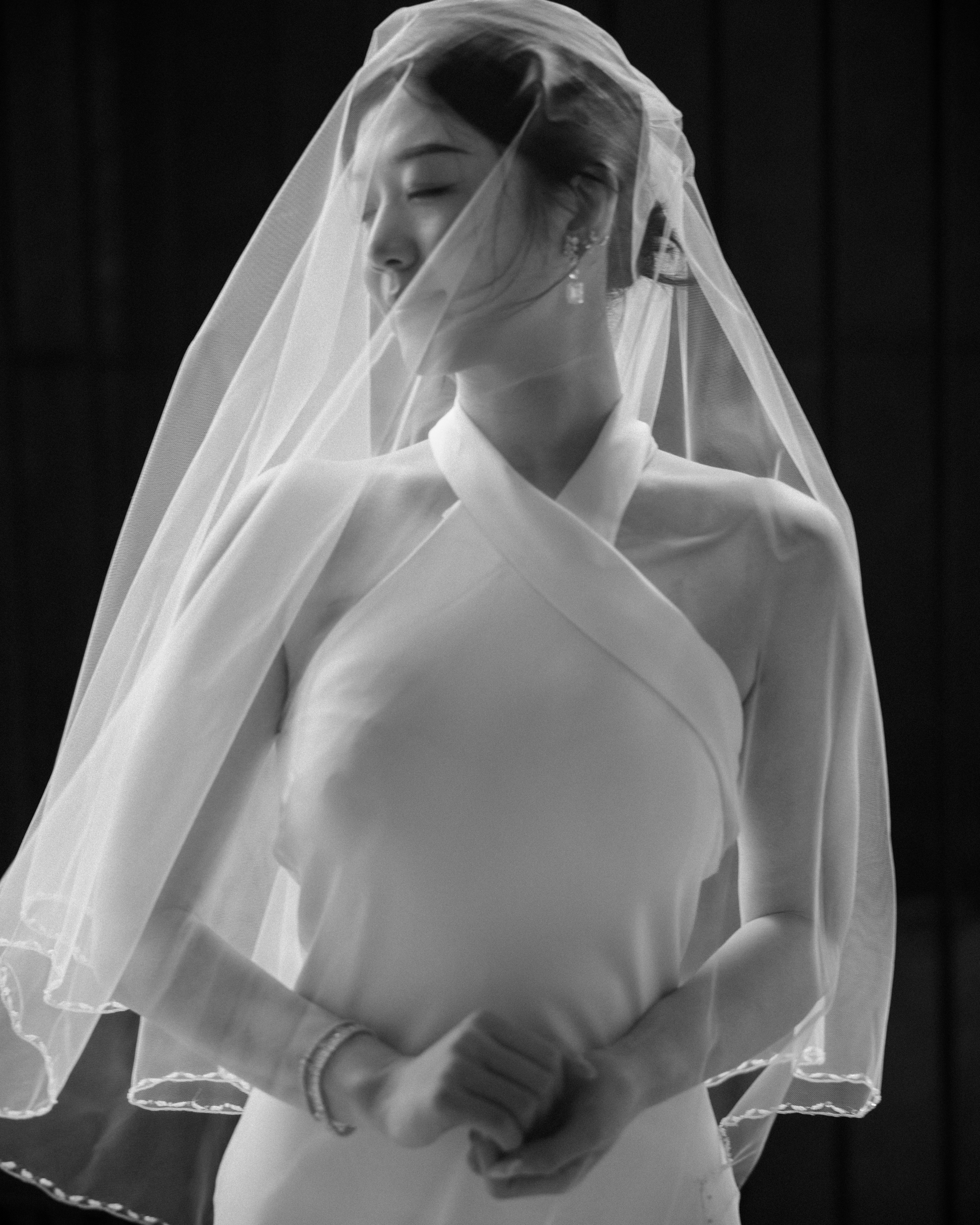 A woman in a sleeveless wedding dress and veil stands with her hands clasped, eyes closed, at the City Hall subway station. The black and white image captures an intimate moment from unique wedding photos.