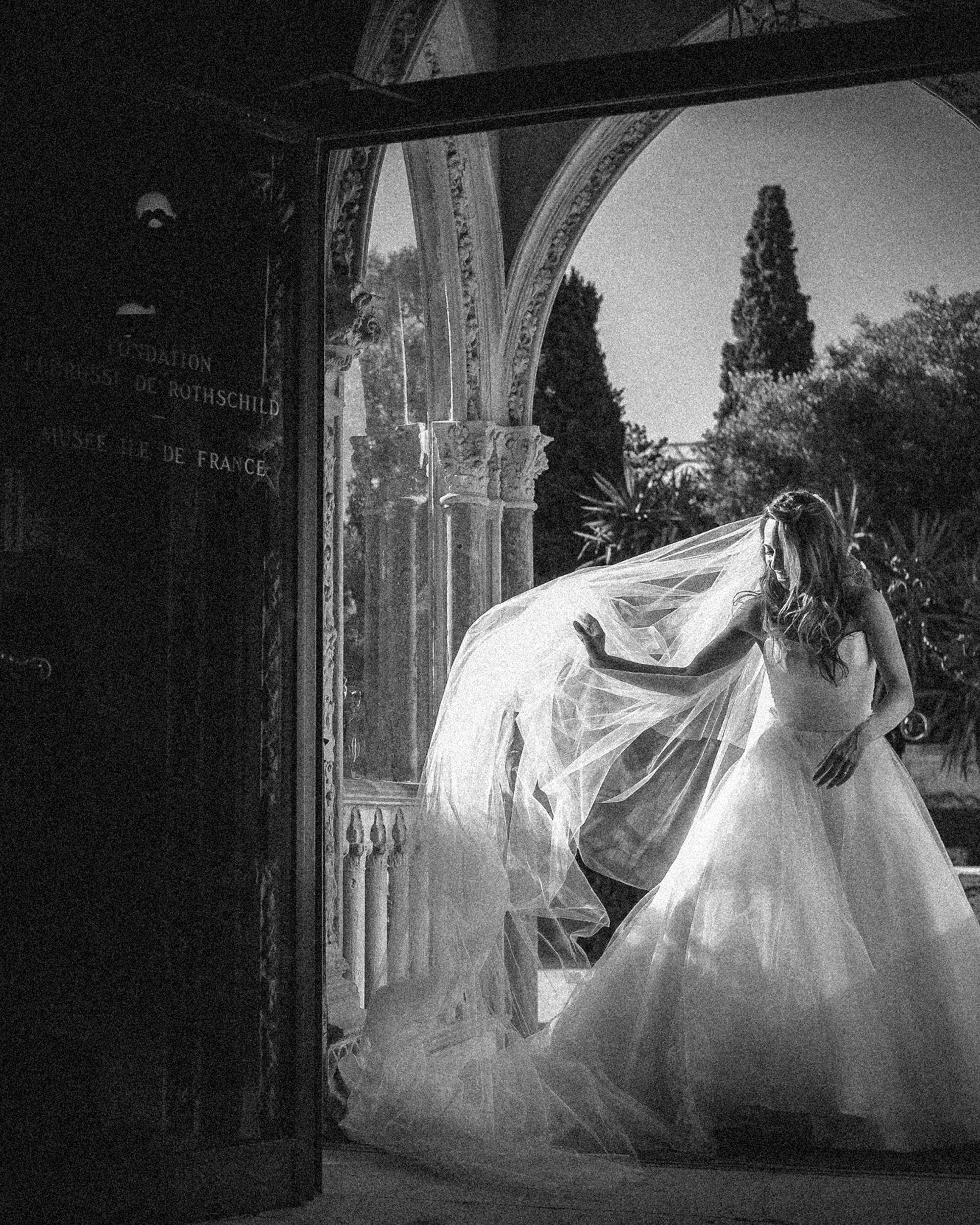 A bride in a flowing wedding gown stands near an arched doorway at her Villa Ephrussi de Rothschild wedding, holding her veil as sunlight streams in from outside. The scene is in black and white.