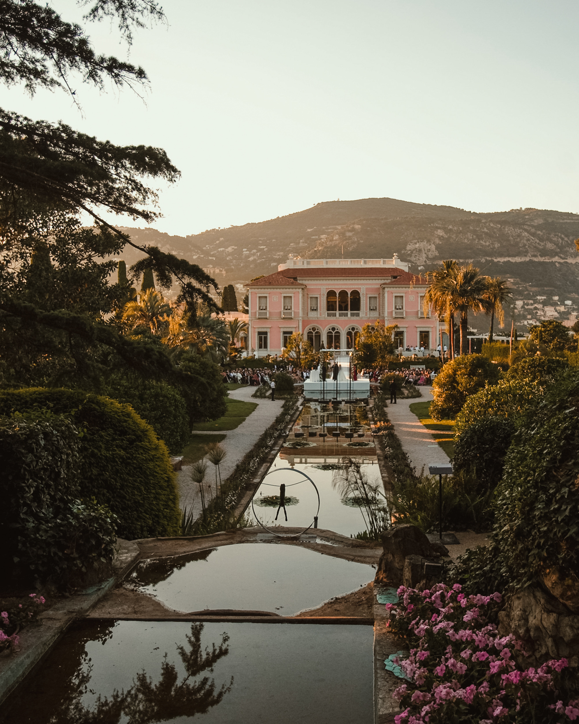 A large pink villa with arched windows stands at the end of a symmetrical garden—an idyllic setting for a luxury destination wedding, with fountains, pools, palm trees, and mountains glowing in the sunset.