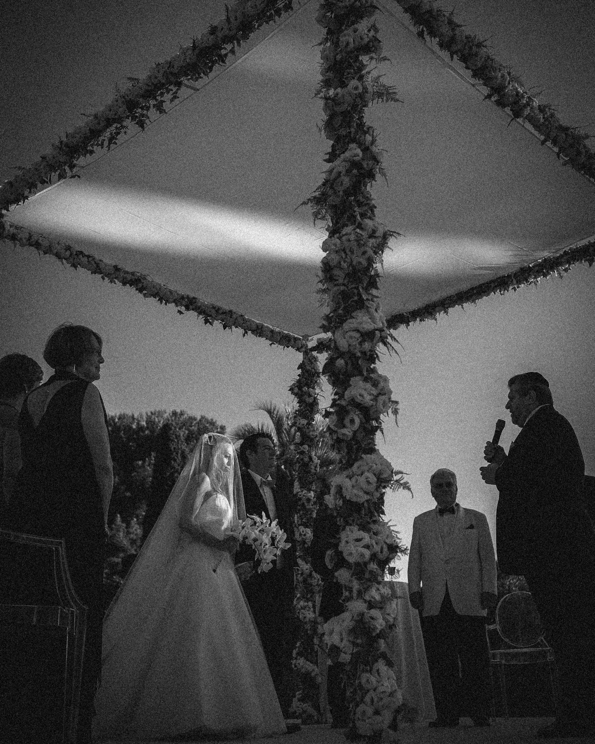 A bride and groom stand under a floral canopy with several people during an outdoor Villa Ephrussi de Rothschild wedding ceremony; one man speaks into a microphone.