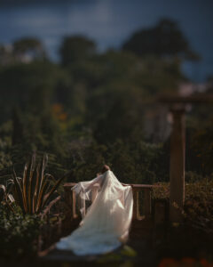 A person in a long white wedding gown and veil stands on a stone terrace at the Villa Ephrussi de Rothschild, surrounded by greenery, gazing out at a blurred landscape—capturing the magic of a destination wedding.