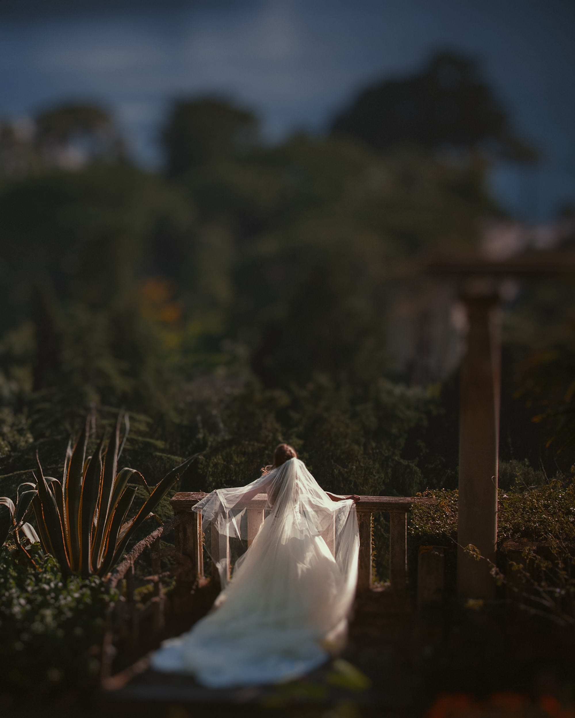 A person in a long white wedding gown and veil stands on a stone terrace at the Villa Ephrussi de Rothschild, surrounded by greenery, gazing out at a blurred landscape—capturing the magic of a destination wedding.