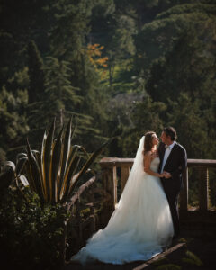 A bride and groom in formal wedding attire stand on a stone balcony at their Villa Ephrussi de Rothschild destination wedding, surrounded by lush greenery, sharing an intimate moment.