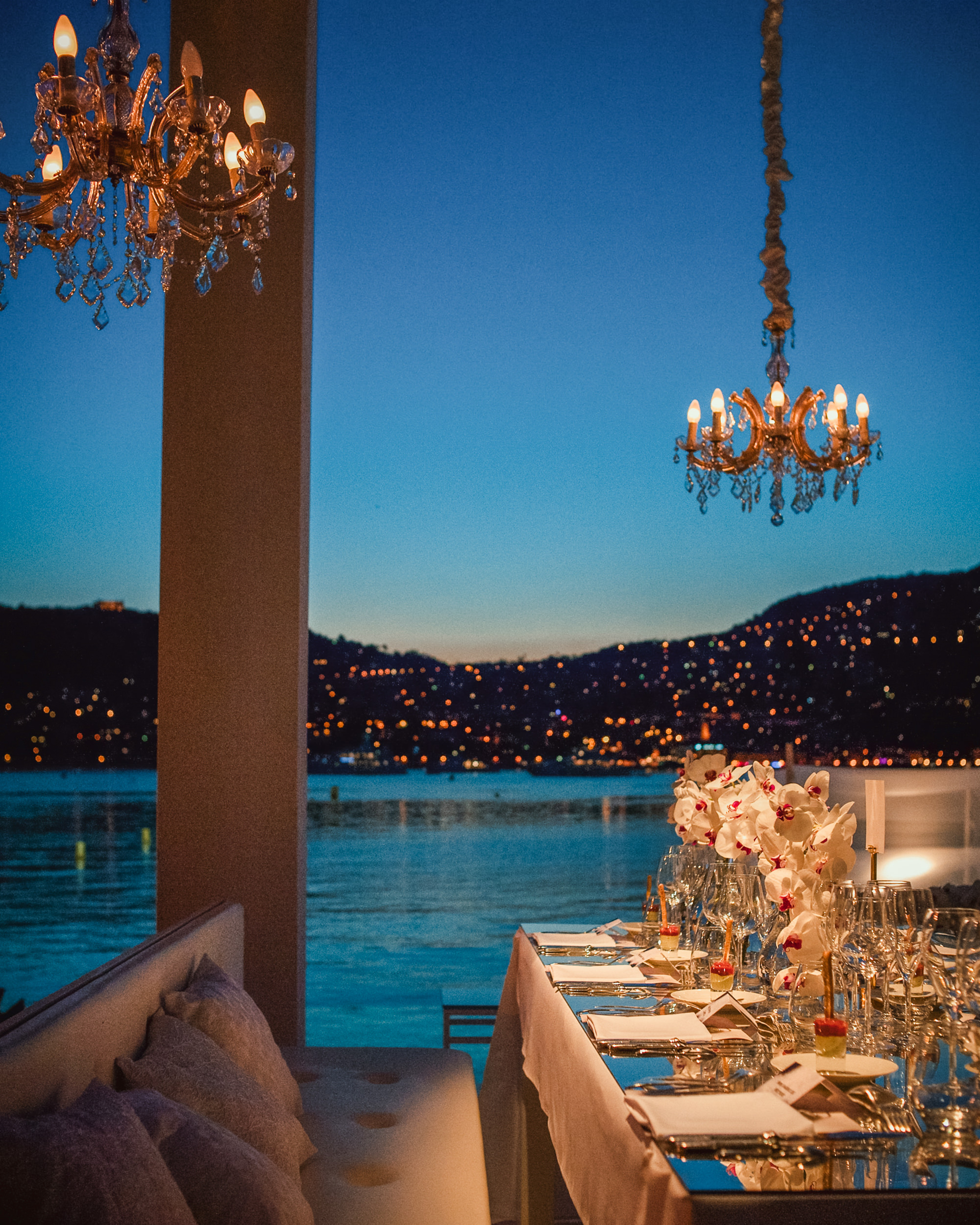 Elegant dinner table set with flowers and glassware beside a lake at dusk, chandeliers overhead and city lights on the hills—perfect for a Luxury Destination Wedding captured by an expert wedding photographer.