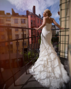 A woman in a white lace wedding dress stands on a city balcony, looking out over colorful apartment buildings under a partly cloudy sky, capturing the charm of a Finland destination wedding.
