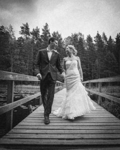 A couple in wedding attire walks hand in hand on a wooden dock surrounded by trees, captured in black and white—an intimate moment from a Finland destination wedding.