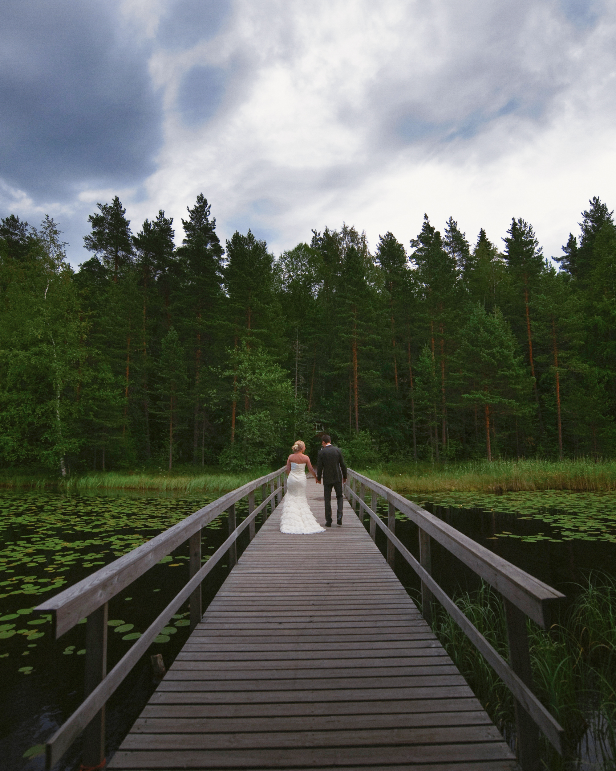 A bride and groom walk hand in hand across a wooden bridge over a pond, surrounded by trees and lily pads under a cloudy sky—a perfect Finland destination wedding setting.