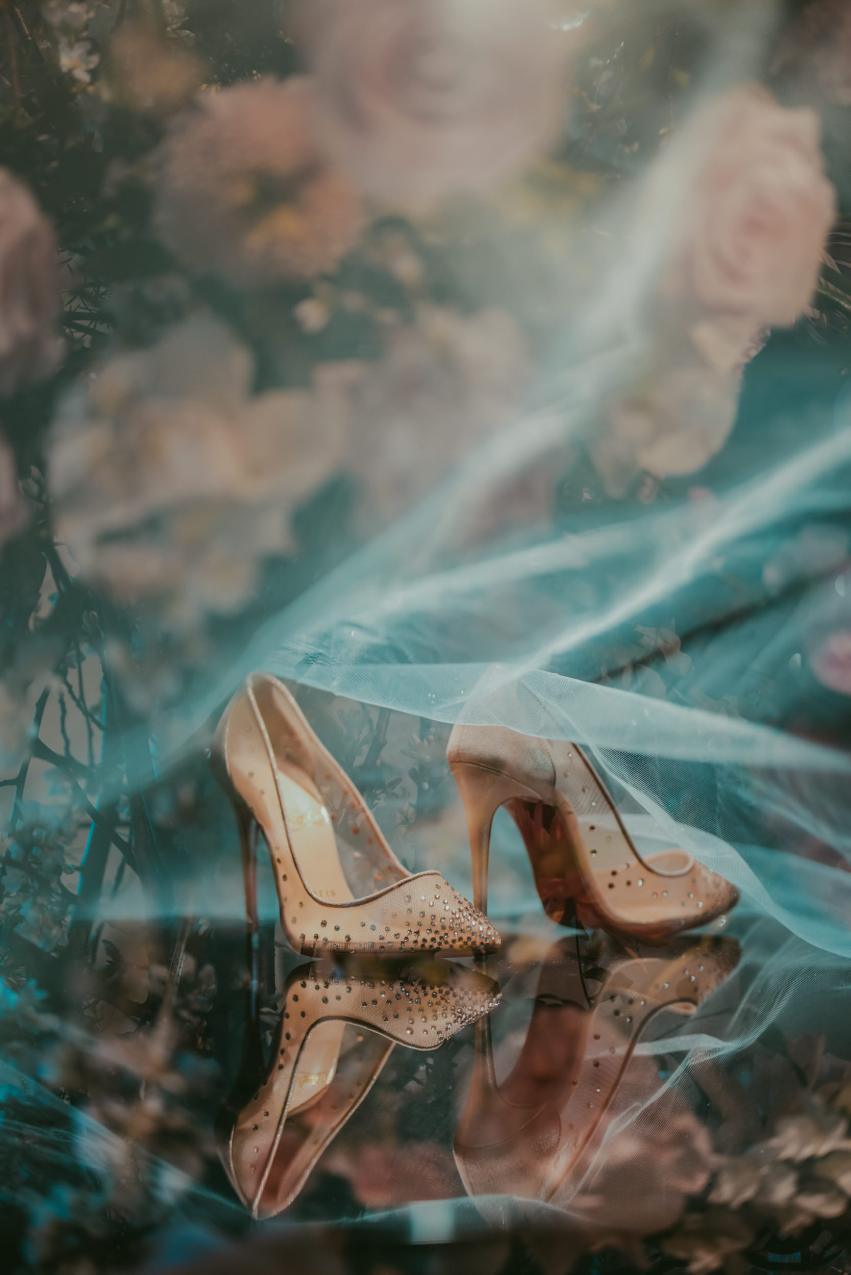 A pair of beige high-heeled shoes with studs, reflected on a surface, surrounded by soft-focus flowers and sheer fabric—perfect elegance for a Liberty Warehouse wedding.