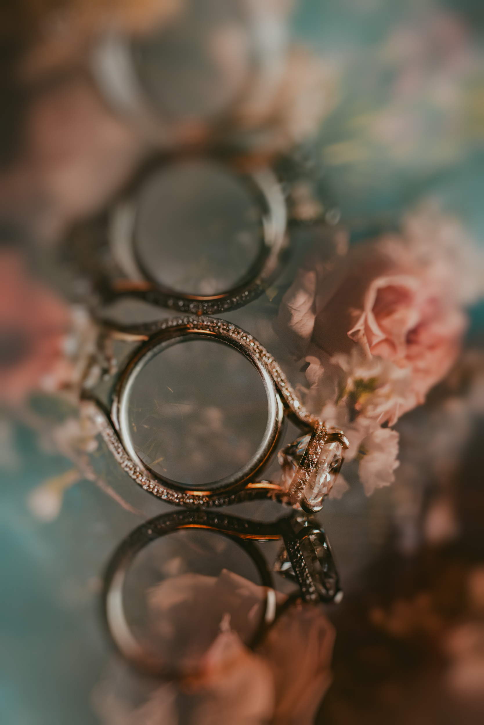 A diamond engagement ring with a decorative band is placed on a reflective surface surrounded by soft pink flowers, creating a dreamy, blurred effect reminiscent of a romantic Liberty Warehouse wedding.