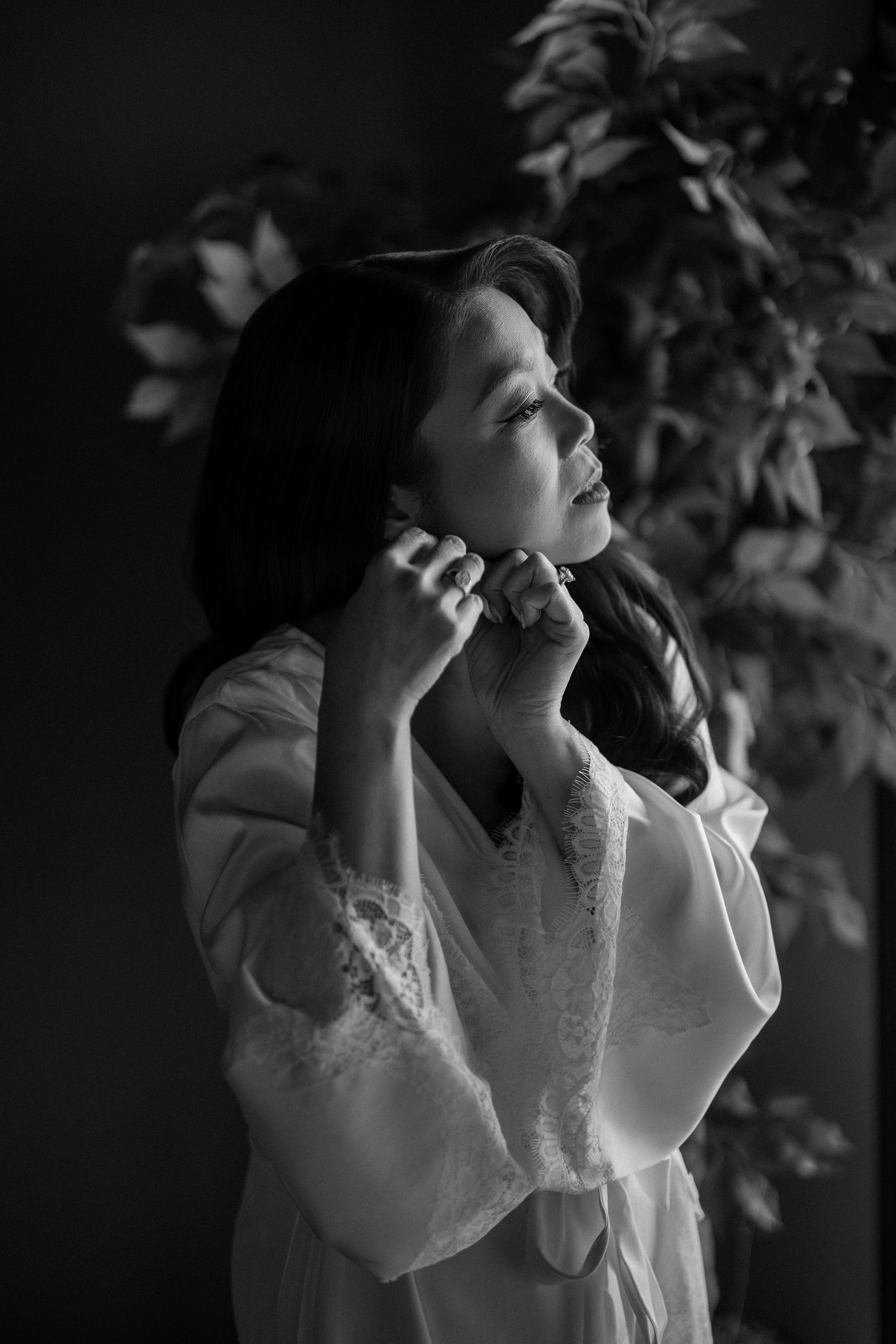 A bride in a robe stands by a leafy plant, putting on an earring, in a softly lit black and white setting reminiscent of a Liberty Warehouse wedding.