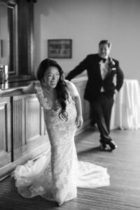 A bride in a lace wedding dress leans forward laughing at her liberty warehouse wedding, while a groom in a tuxedo stands in the background near a bar, smiling.