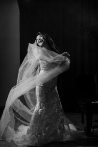 A bride in a lace wedding dress and veil stands beside a piano at her Liberty Warehouse wedding, turning with a joyful expression. The image is in black and white.