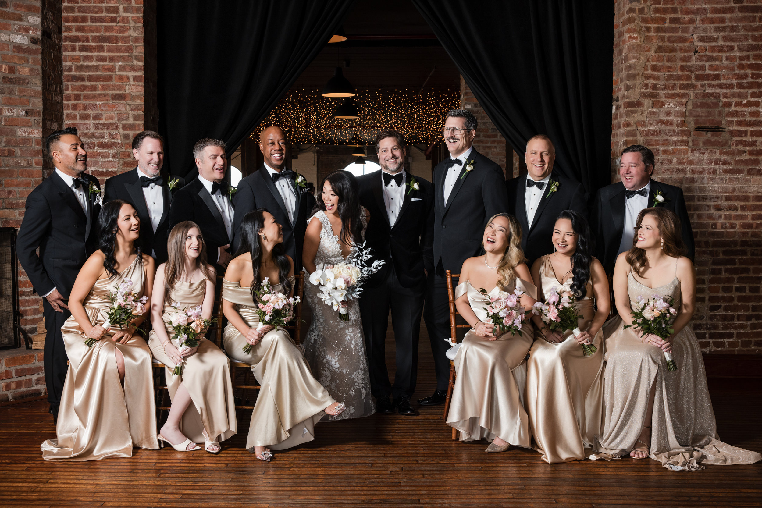 A wedding party poses indoors against a brick wall at a Liberty Warehouse wedding. The group includes six bridesmaids in beige dresses and seven groomsmen in black suits, with the bride and groom standing at the center.