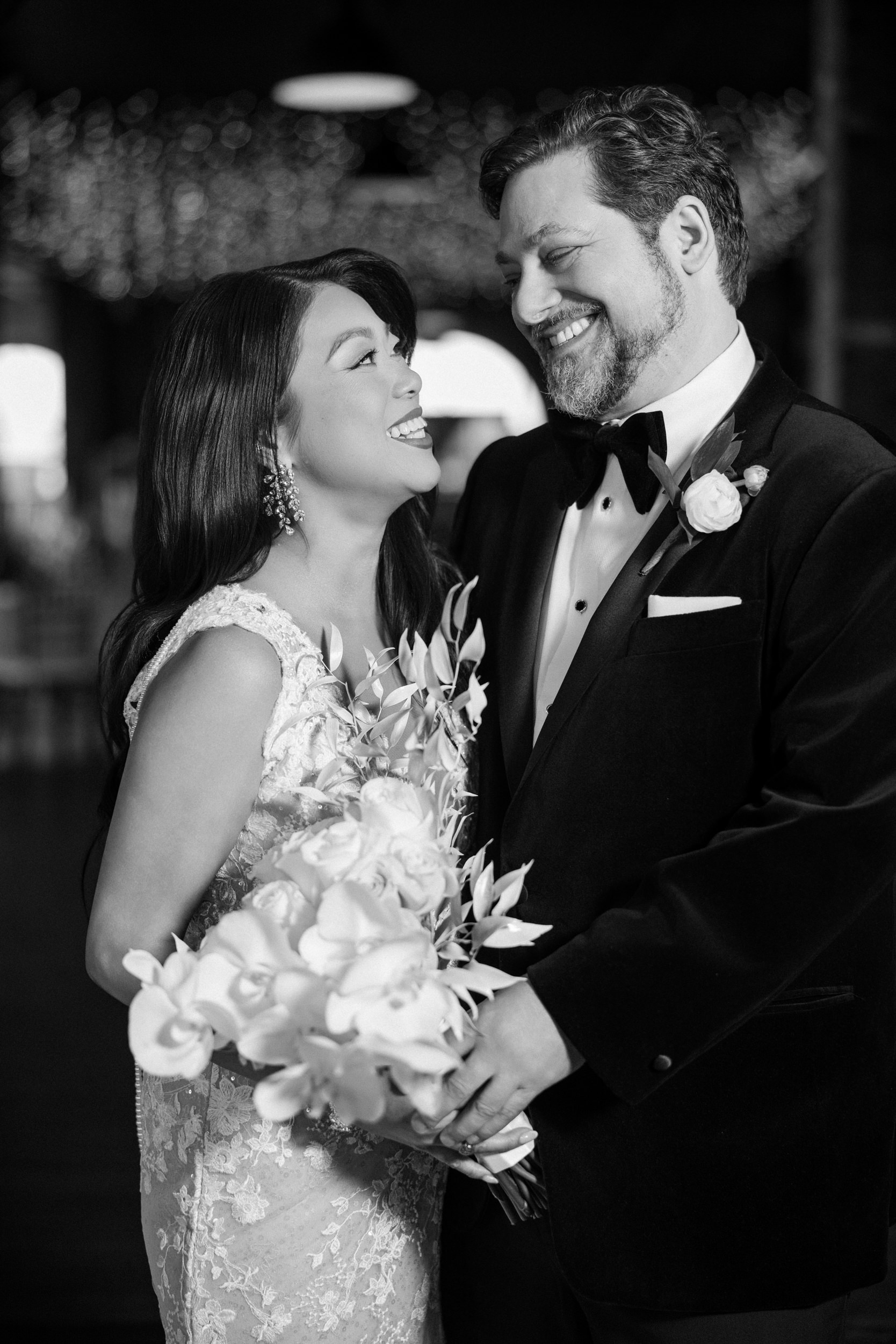 A bride and groom in formal attire stand close together, smiling at each other during their Liberty Warehouse wedding. The bride holds a bouquet of flowers. The image is in black and white.