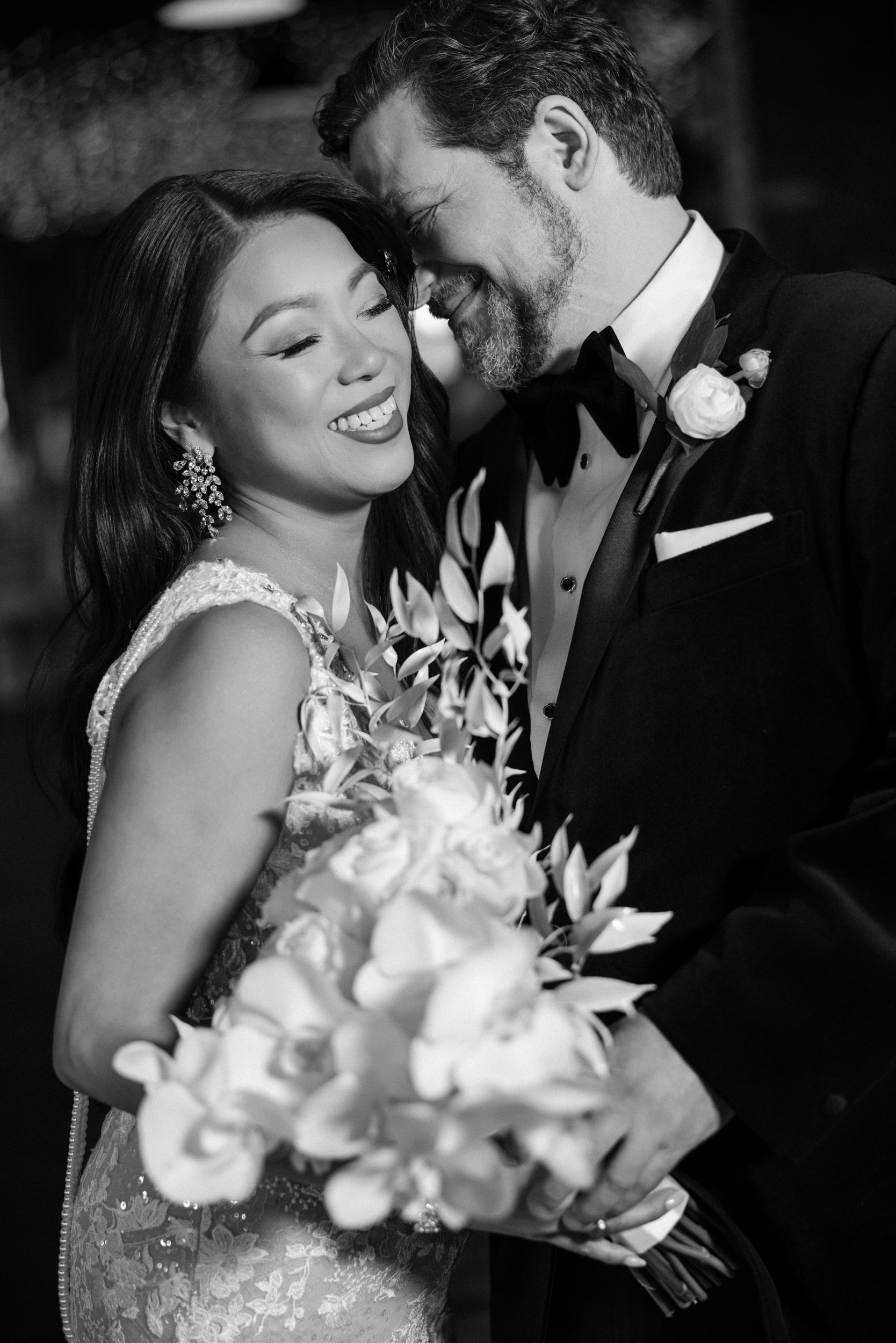 A bride and groom in formal attire smile and embrace, holding a bouquet of flowers, in a black-and-white photo from their unforgettable Liberty Warehouse wedding.
