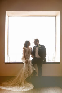 A bride in a lace wedding dress and a groom in a tuxedo sit on a window ledge at their Liberty Warehouse wedding, facing each other as bright light streams in from behind.