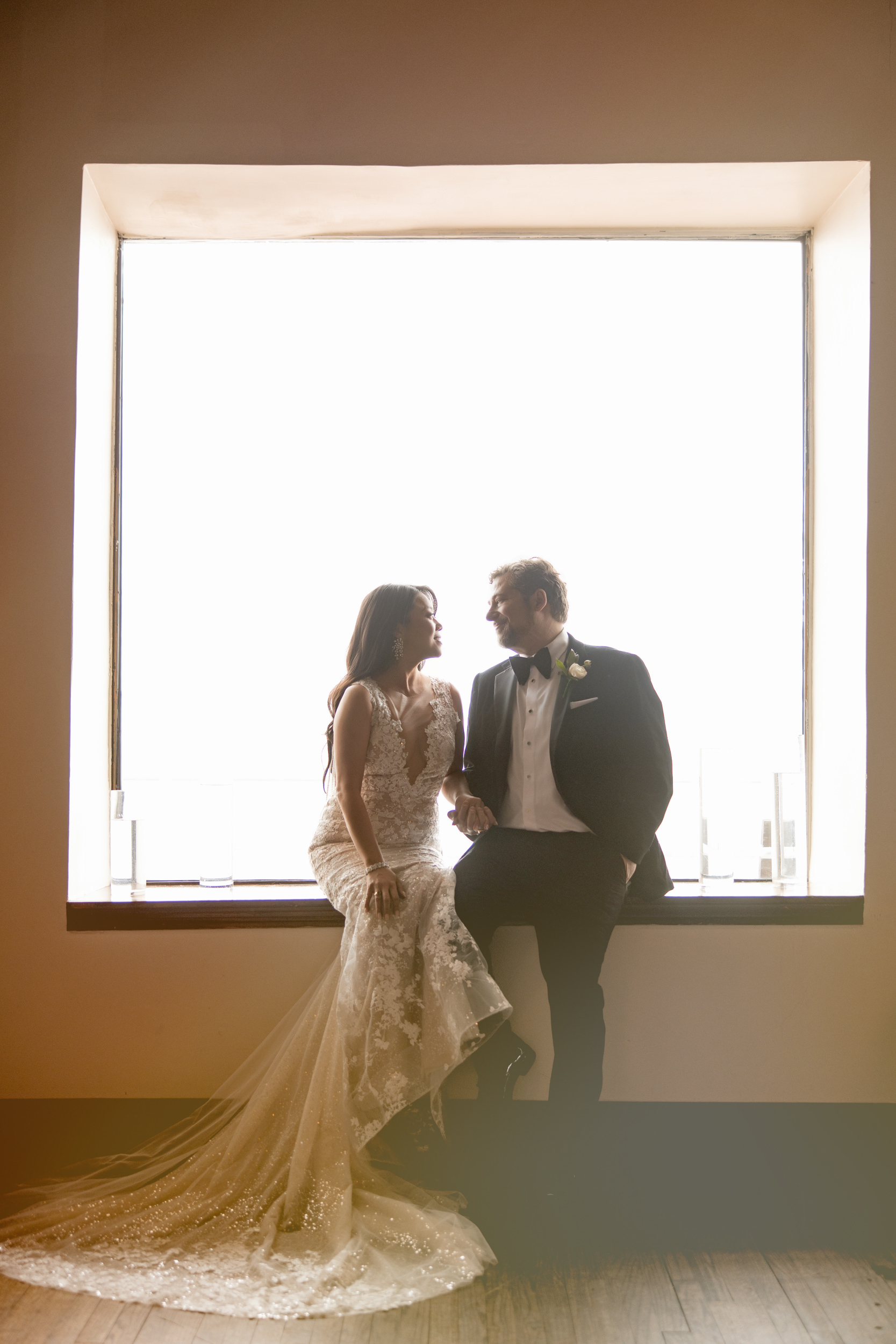 A bride in a lace wedding dress and a groom in a tuxedo sit on a window ledge at their Liberty Warehouse wedding, facing each other as bright light streams in from behind.