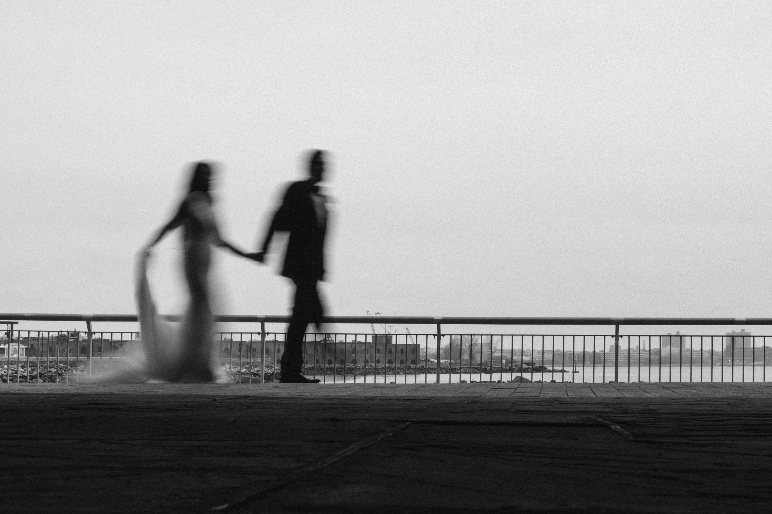 A blurred black-and-white image shows a couple in wedding attire holding hands and walking outdoors near a railing by the water, capturing the romantic ambiance of a Liberty Warehouse wedding.