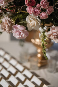 A gold vase holds pink and white roses above a table with rows of place cards and glass votive candle holders at a stunning Liberty Warehouse wedding.