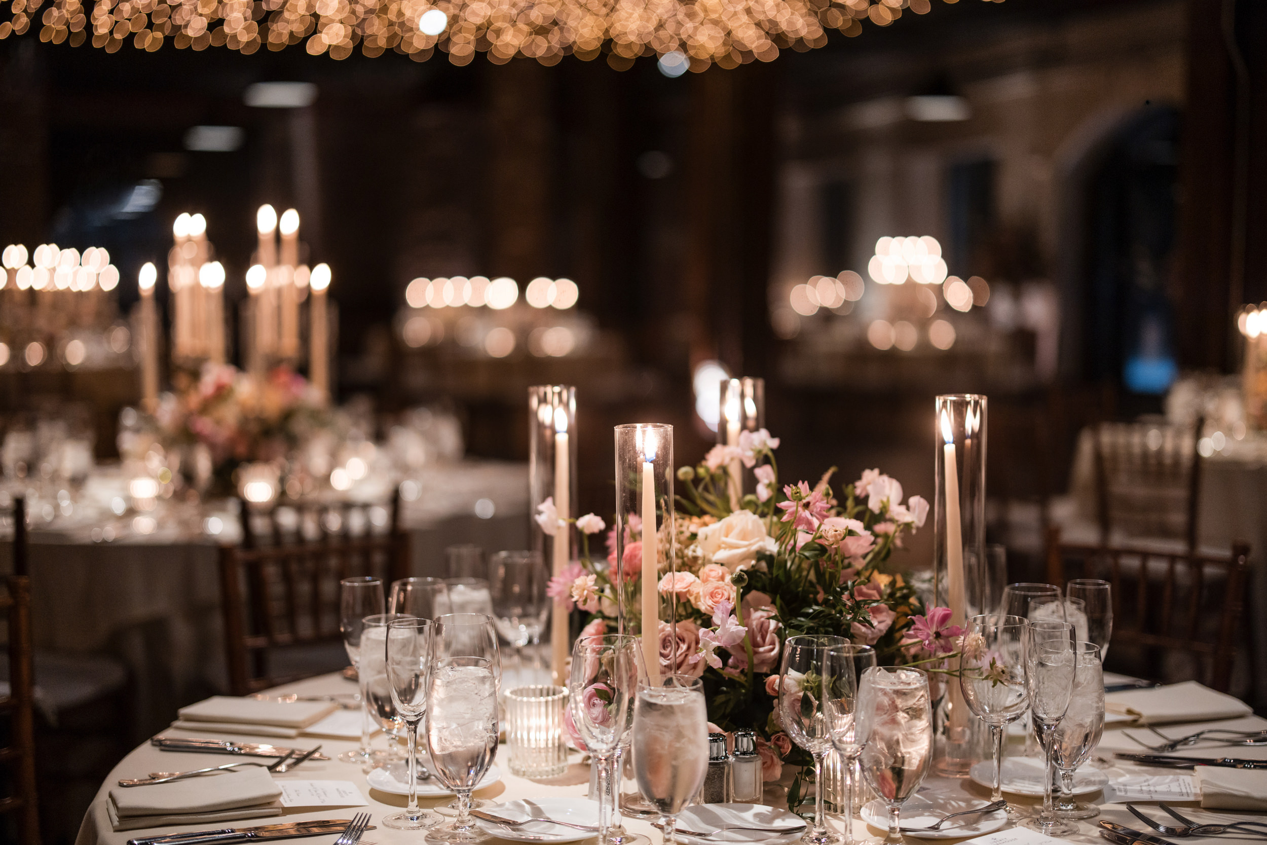 Elegant banquet tables set with glassware, floral centerpieces, and tall candles under string lights in a dimly lit Liberty Warehouse wedding hall.