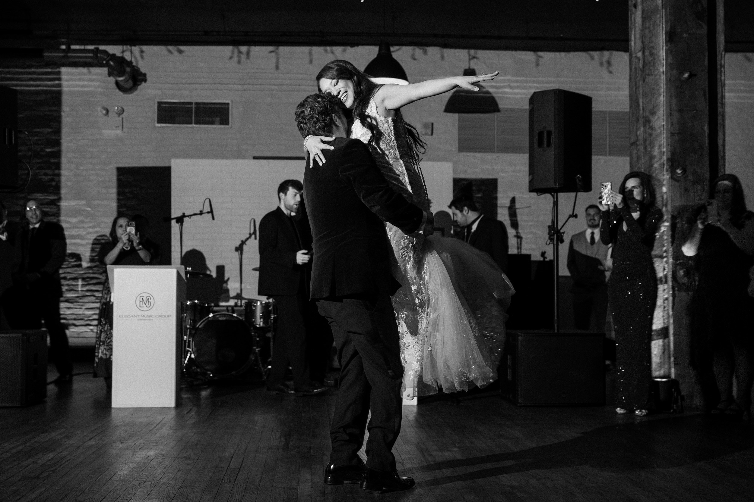 A man lifts a woman in a dress as they dance on the wooden floor at a formal event in Liberty Warehouse, with a band and guests watching in the background.