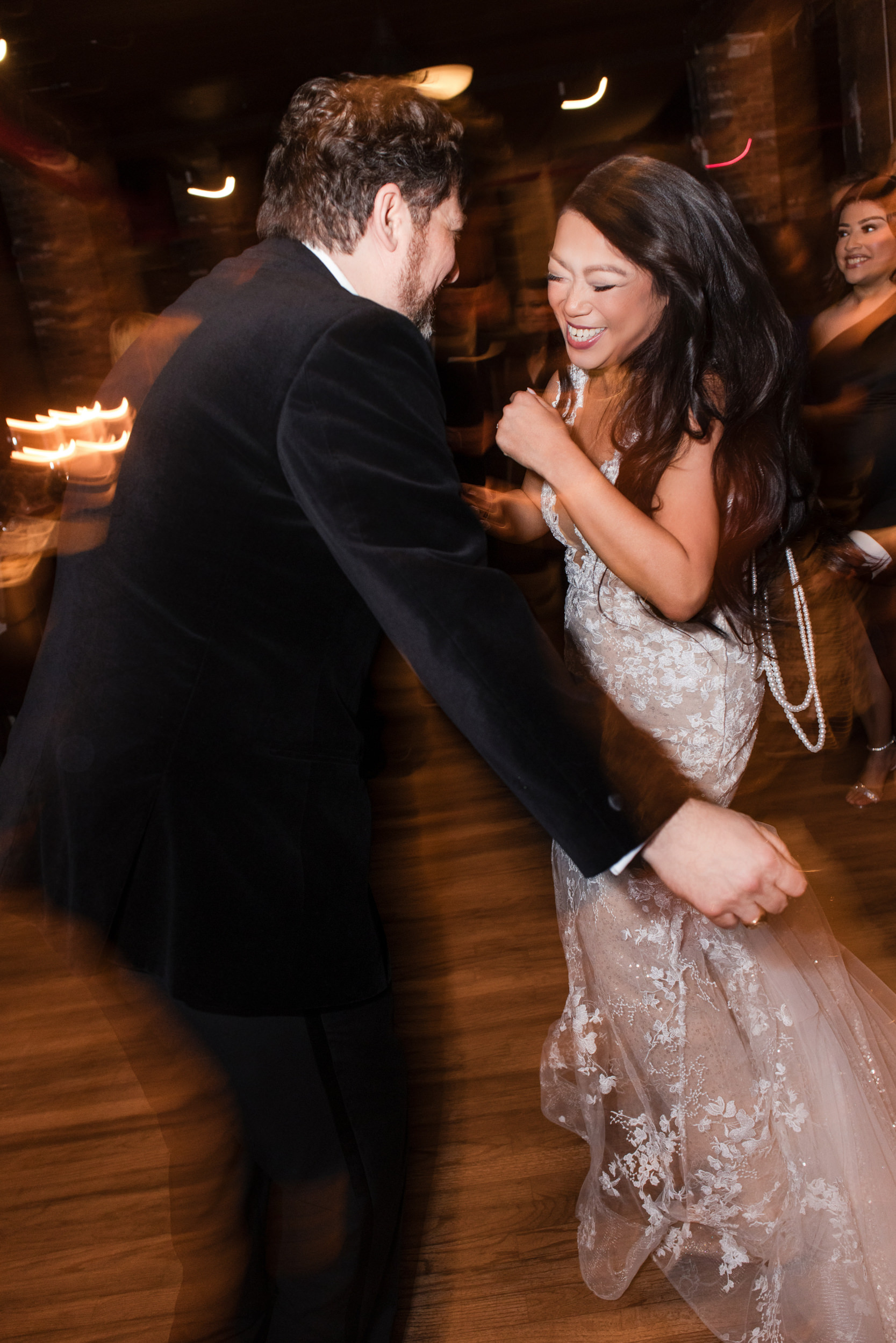 A man in a black suit and a woman in a white lace dress are dancing together indoors at their Liberty Warehouse wedding, smiling and holding hands.