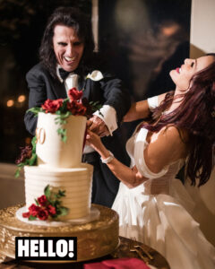 Alice Cooper in a tuxedo and a Sheryl Cooper in a white dress cut a two-tiered cake decorated with red roses, both smiling and laughing at their wedding.