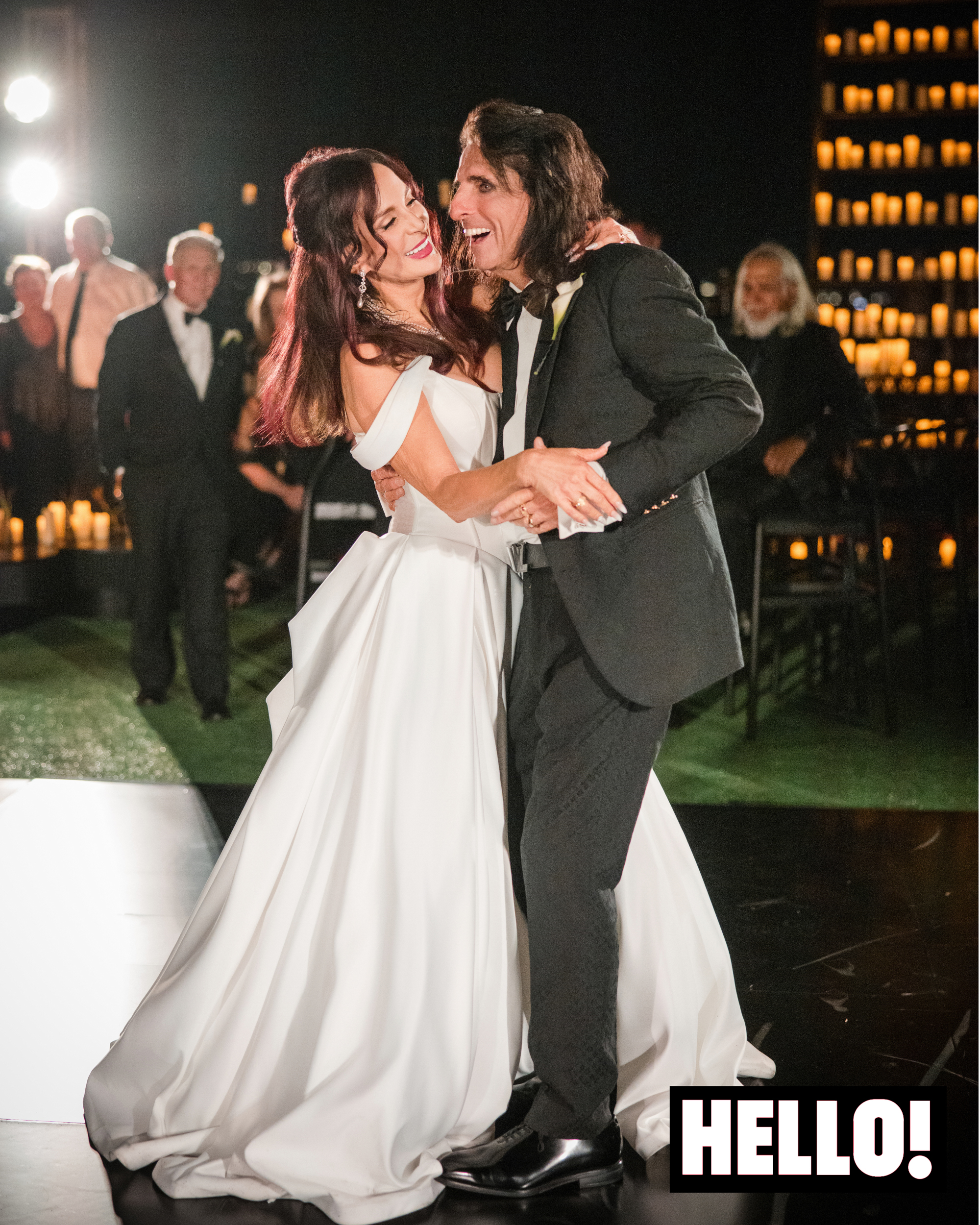Alice Cooper and Sheryl Cooper, Alice in a Louis Vuitton suit and Sheryl in a bridal gown, smile and dance together at their wedding reception at Wrigley Mansion with guests in the background.