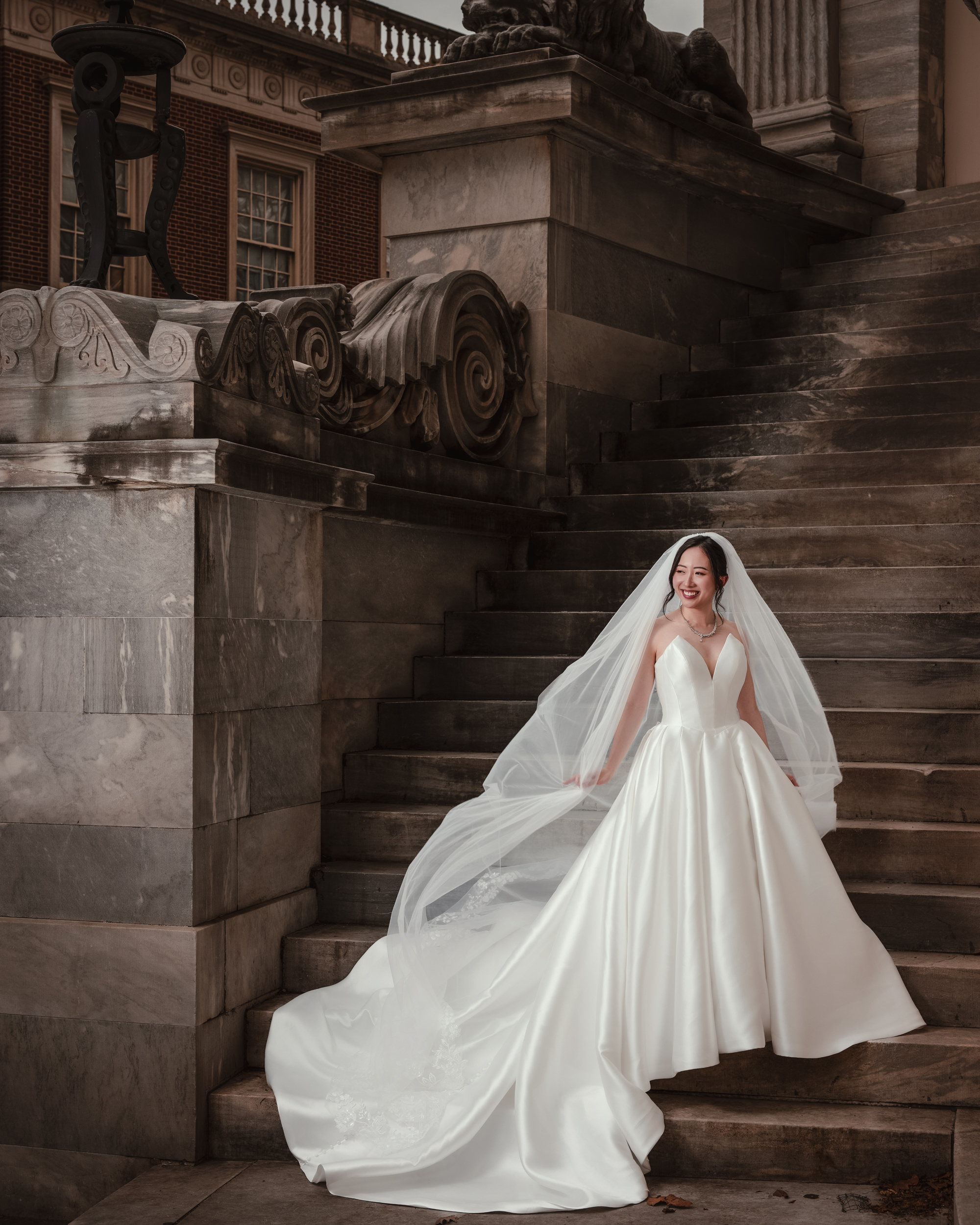 A bride in a white wedding dress and veil stands on stone steps beside the ornate Merchant's Exchange, smiling and holding her dress for elegant Downtown Club Philadelphia wedding photos.