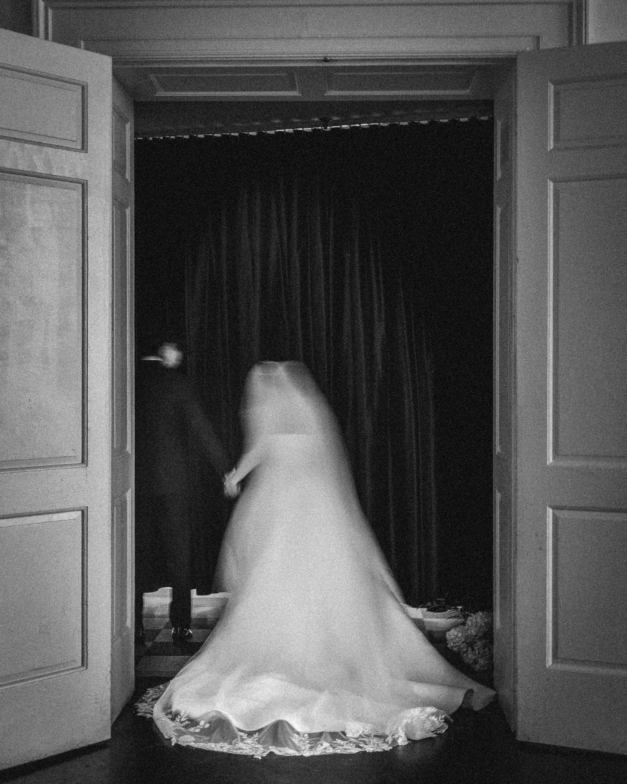 A blurred black and white photo shows a bride and groom holding hands as they walk through double doors toward a dark curtain at their Downtown Club Philadelphia wedding.