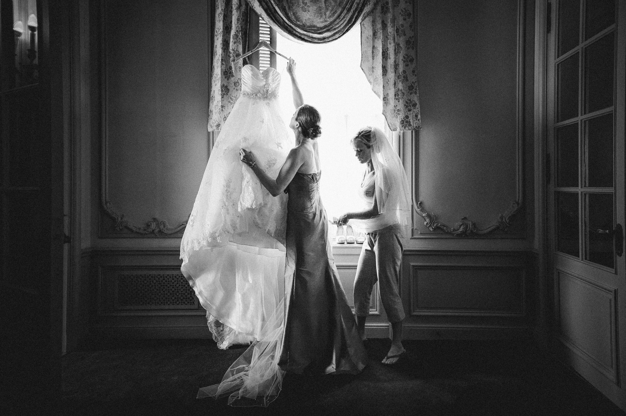 Two women stand by a window at an Elkins Estate wedding; one holds a wedding dress up to the light while the other, wearing a veil, looks on in the ornate room.