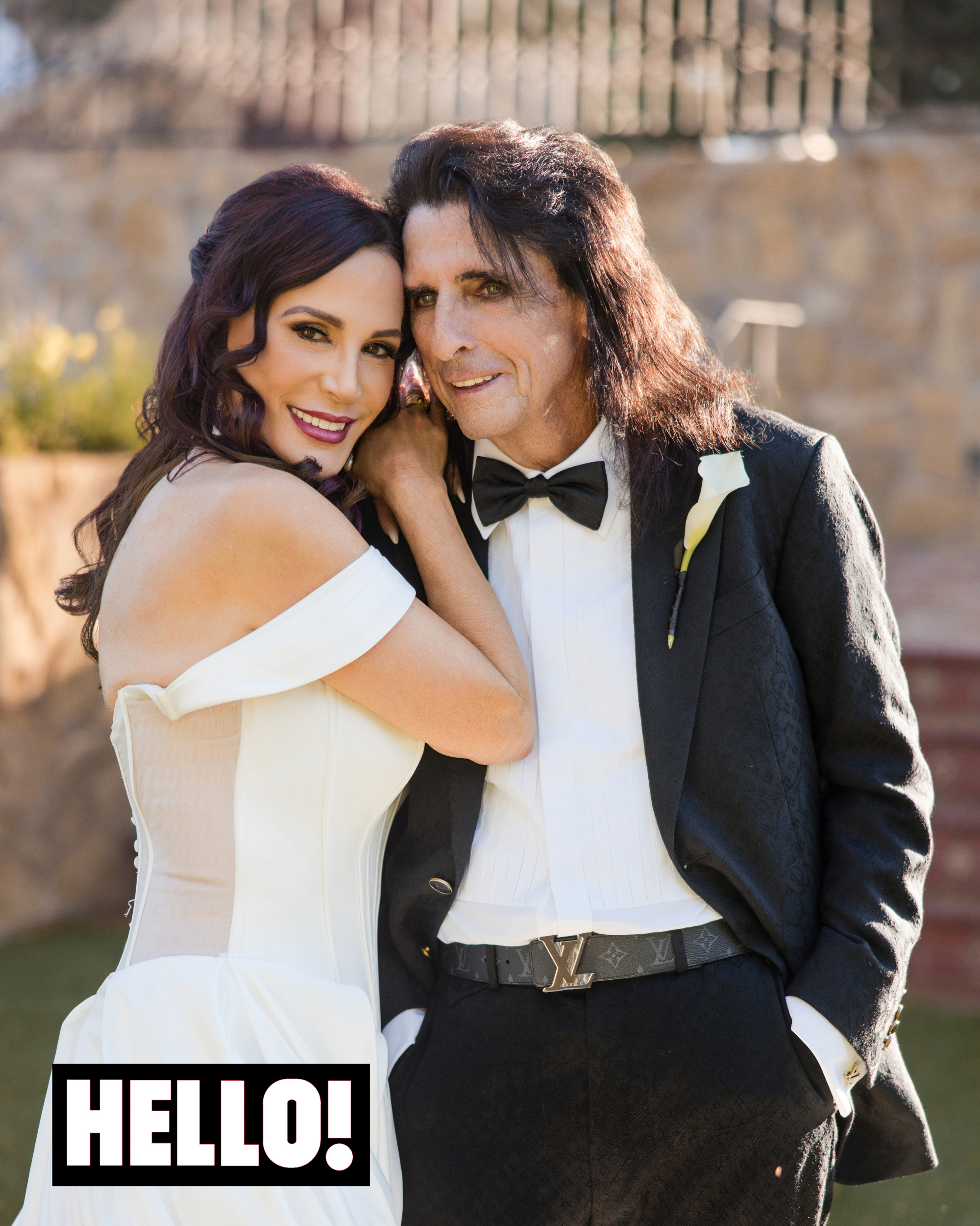 Alice Cooper and Sheryl Cooper, dressed in wedding attire pose closely outdoors at Wrigley Mansion; Sheryl wears a white gown and Alice wears a tuxedo with a bow tie.
