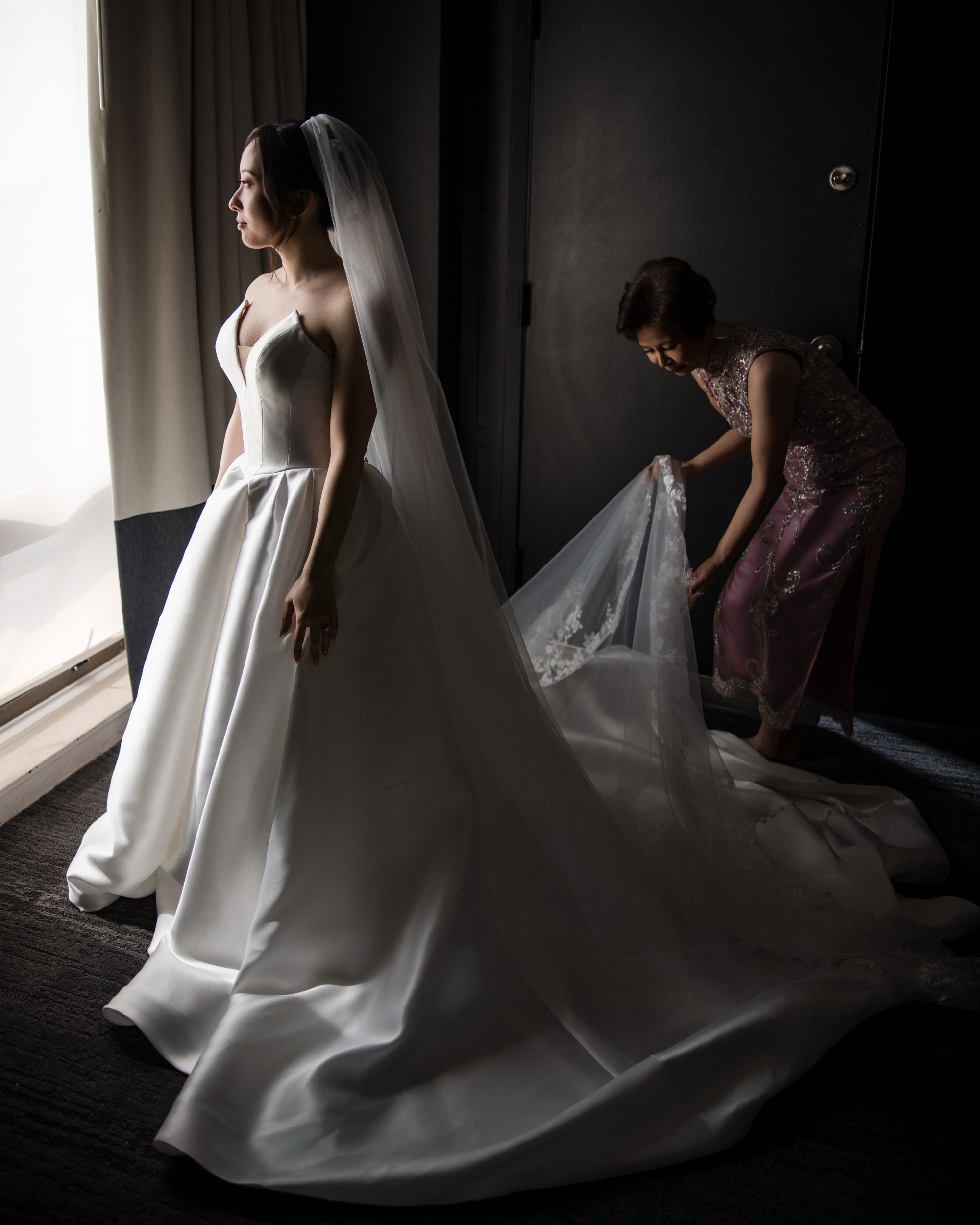 A bride in a white gown stands by a window at the Downtown Club Philadelphia Wedding, looking out, while another woman arranges the train of her dress.
