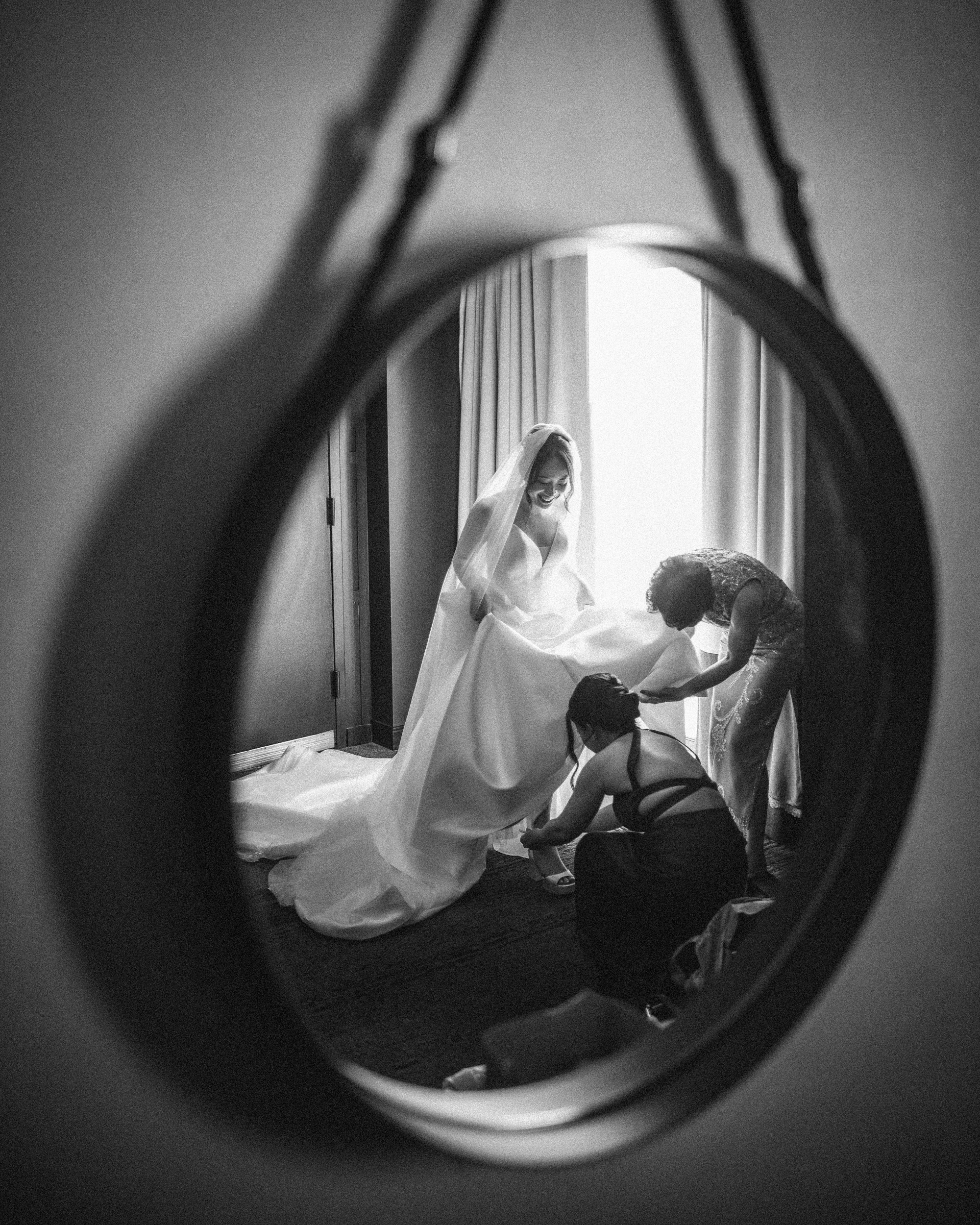 Black and white photo of a bride in a wedding dress being assisted by two people, seen through the reflection of a round mirror on her Downtown Club Philadelphia Wedding day.