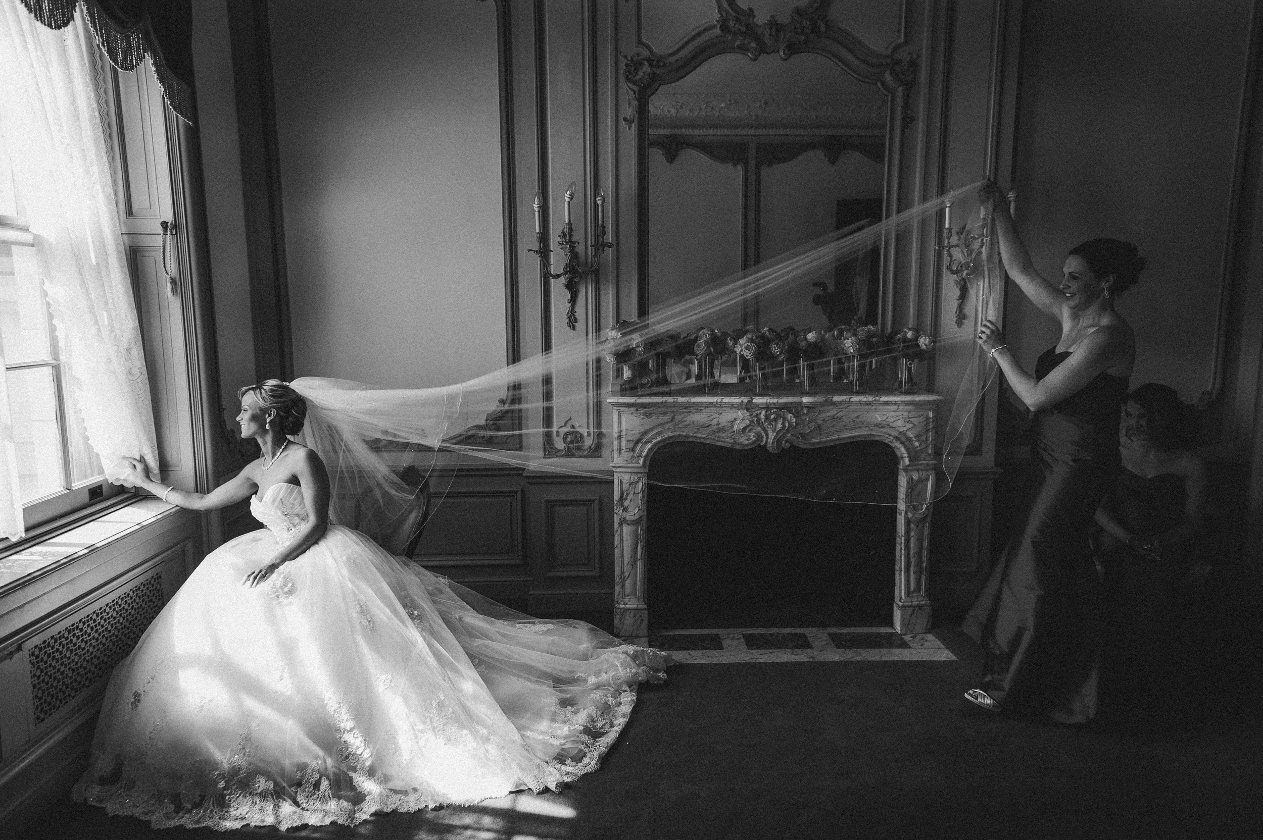 A bride sits by a window in a formal room during an Elkins Estate wedding while another woman holds her veil, stretched out near a fireplace with a large mirror above it. A bride sits by a window in a formal room during an Elkins Estate wedding while another woman holds her veil, stretched out near a fireplace with a large mirror above it.