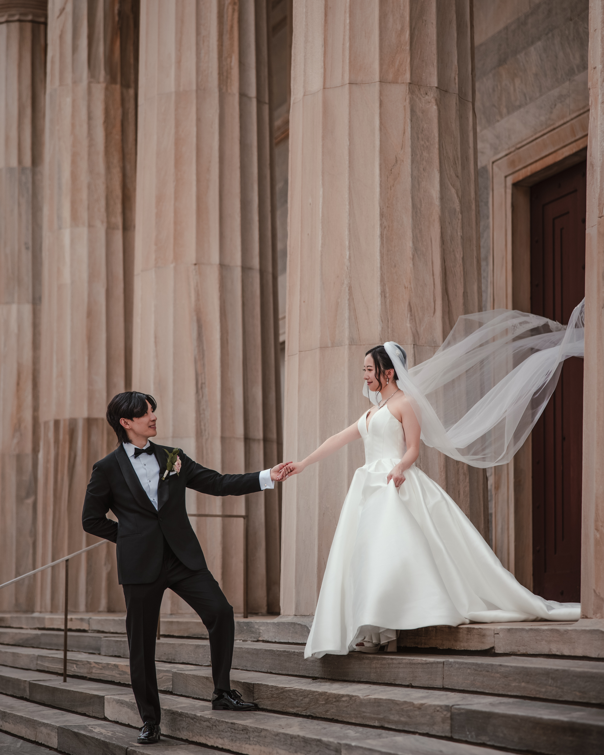 A bride in a white gown and veil stands on steps, holding hands with a groom in a black tuxedo, captured during their Downtown Club Philadelphia wedding second bank photos.
