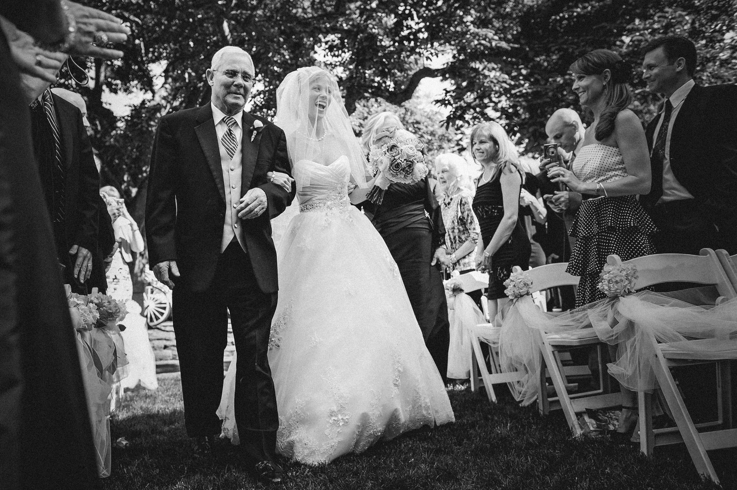 At an Elkins Estate wedding, a bride in a wedding gown walks down the aisle outdoors, escorted by an older man, as guests watch and smile. At an Elkins Estate wedding, a bride in a wedding gown walks down the aisle outdoors, escorted by an older man, as guests watch and smile.
