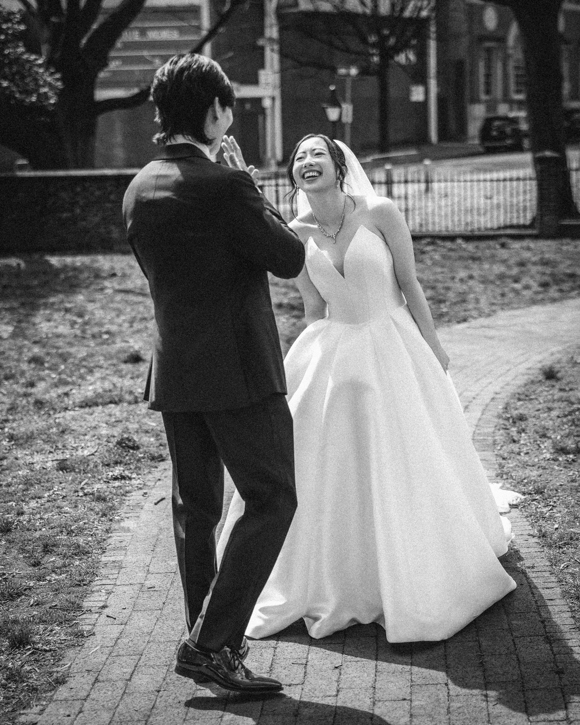 A bride in a white gown and a groom in a dark suit stand outside on a pathway, smiling and laughing together after their Downtown Club Philadelphia Wedding.